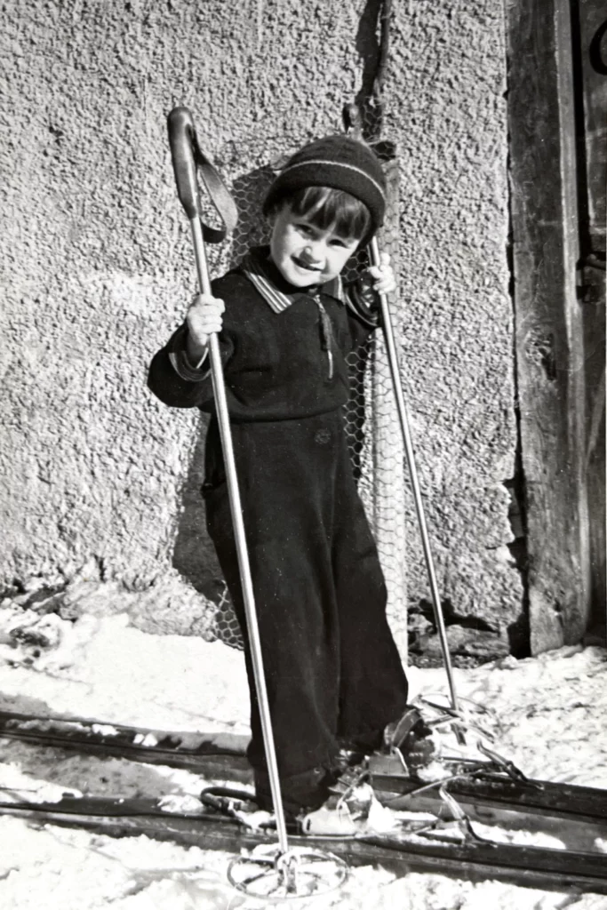 Pierrot Michaud enfant, à skis devant un mur de chalet, ses bâtons à la main.
